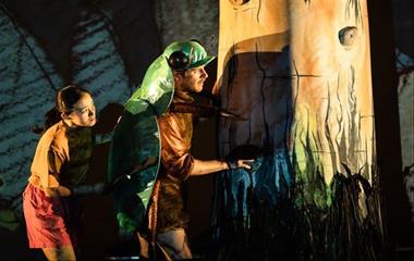 Two performers wearing costumes on stage standing next to a tree.