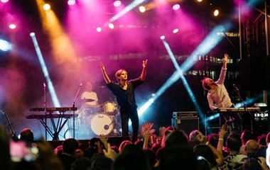 Three artists on stage with microphones and instruments performing to a crowd.