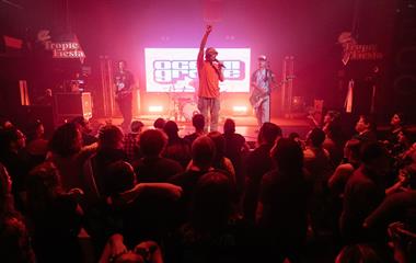 Three artists on stage with microphones and instruments performing to a crowd.